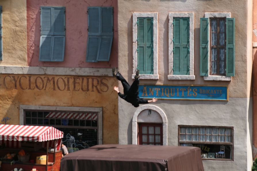 A stunt performer mid-leap on a European-style street set at a theme park show